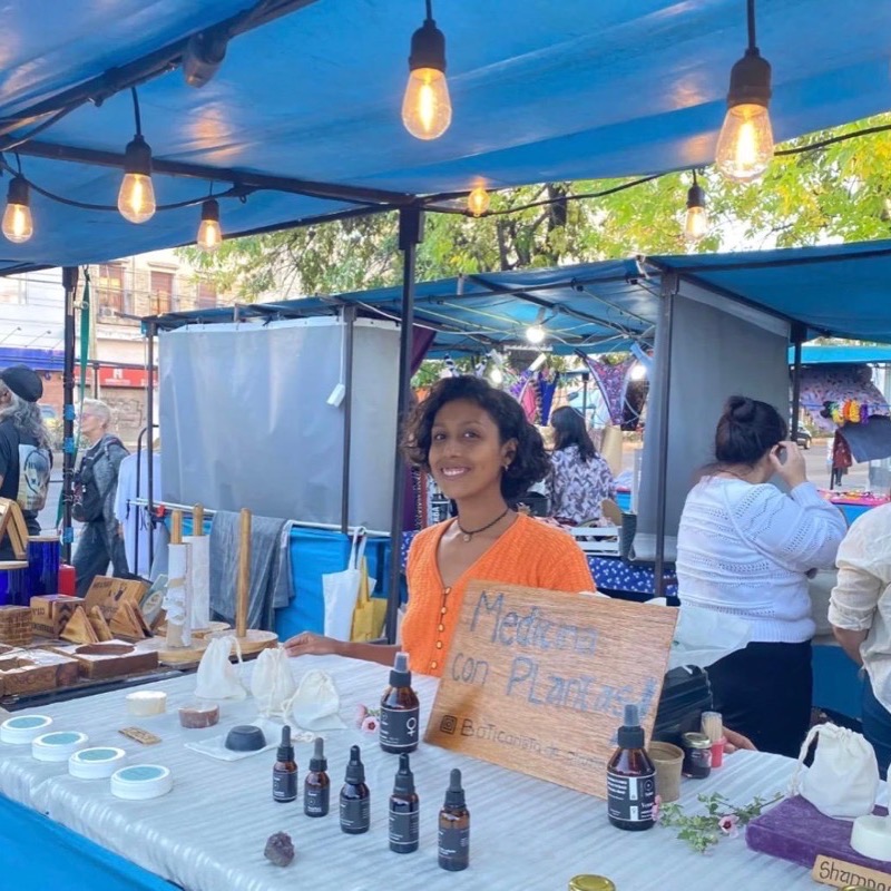 Jenny en su puesto de feria, con sus frascos y un cartel escrito a mano que dice Medicina con Plantas